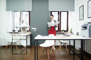 Confident young businesswoman in eyeglasses leaning to wall while doing paperwork, interior of modern open plan office on background