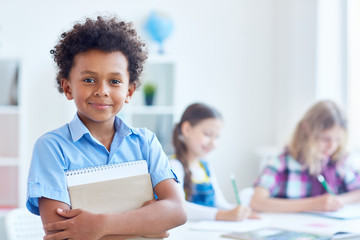 Fototapeta premium Cute schoolboy with copybook looking at camera with his classmates on background