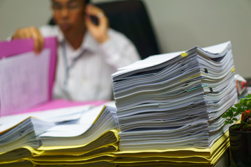 Pile of organized but unfinished business documents on desk with businessman talking on  smartphone and sitting on the chair in the office background,  business and busy job concept