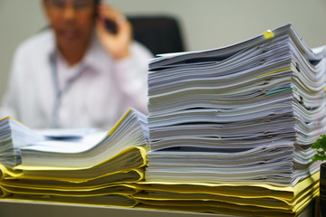 Pile of organized but unfinished business documents on desk with businessman talking on  smartphone and sitting on the chair in the office background,  business and busy job concept