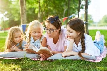 Teacher and three little girls reading stories from book while relaxing on grass