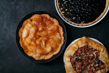 Variety of autumn pies: blueberry tart, chantarelle galette and classical apple pie with sweet pastry crust. Harvest time baking concept. Dark rustic background. Space for text.