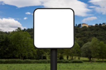 Square white sign with rounded corners. In the background a park and a hill with houses. The background is blurry.