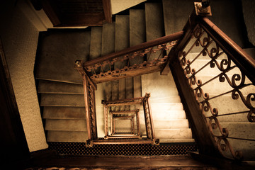 Old and dark squared spiral multi-flight stairway with brown wood and metal handrails seen from the top of the staircase.