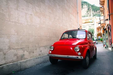  Red old well-preserved vintage Italian classic car parked in a small alley in an Italian Sicilian city with a large empty stone wall in the left side of the frame. © fewerton