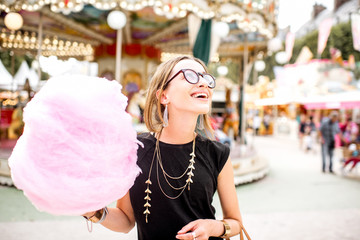 Fototapeta premium Young woman standing with pink cotton candy outdoors in front of the carrousel at the amusement park
