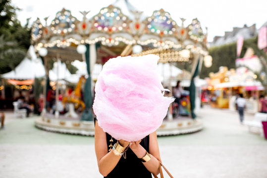 Young woman standing with pink cotton candy outdoors in front of the carrousel at the amusement park