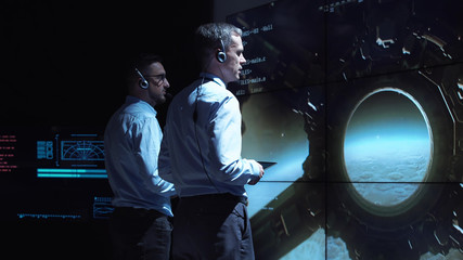 Back view of two supervisors standing at screen in space center and controlling landing on Moon. - obrazy, fototapety, plakaty