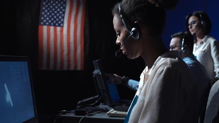 Side view of black woman sitting and working at computer in space mission control center. Elements of this image furnished by NASA. - obrazy, fototapety, plakaty