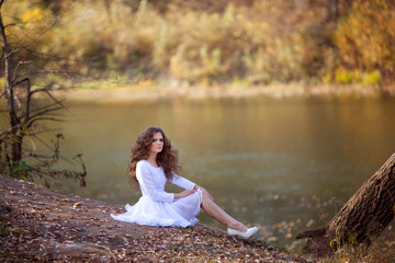 Beautiful young girl of European appearance with makeup and hairstyle, in a white dress, sitting in the autumn forest near the river