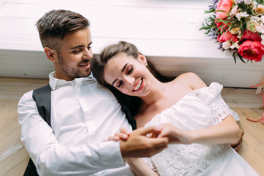 Cheerful newlyweds holding hands and lie on the floor next to bridal bouquet. Artwork. Wedding morning. Soft focus
