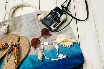 Flat lay of summer fashion with camera, slippers, sunglasses and other girl accessories on top of the bag on white wooden background