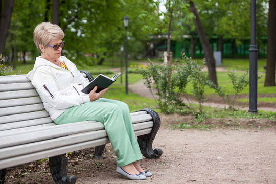 Full length portrait of elder woman in glasses reading book on bench in park, copy space