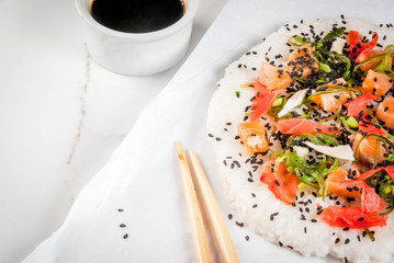 Trend hybrid food. Japanese, Asian cuisine. Sushi pizza with salmon, hayashi wakame, daikon, pickled ginger, red caviar. On a white marble table, with chopsticks and soy sauce. Copy space
