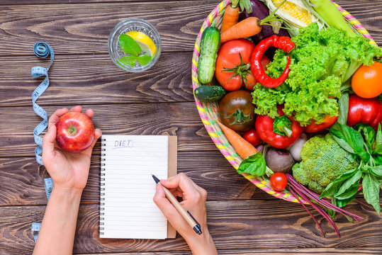 Female hands make notes in diet book on the wood table with Basket of different Fresh Vegetables, measuring tape, glass of water. Healthy lifestyle concept. Detox, diet. Selective focus. Copy space