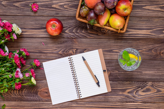 Top view workplace - wooden table with notebook, basket of seasonal fruits, glass of lemon water and Chrysanthemums flowers. Time for planning. Healthy lifestyle concept. Selective focus. Copy space