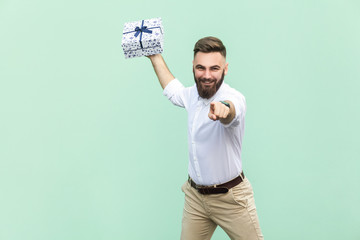 Its for you! Young adult businessman, holding gift box, pointing finger at camera and toothy smile. Isolated on light green background. Indoor, studio shot - obrazy, fototapety, plakaty