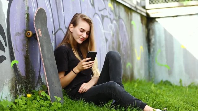 Young girl with skateboard is holding smartphone against the background of graffiti wall