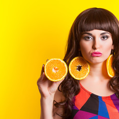 beautiful young woman wearing earrings made from orange and holding orange half against yellow background