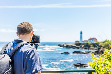 Back of man photographer taking pictures on tripod of Portland Head Lighthouse in Fort Williams park in Maine during summer day