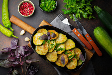 Fried zucchini with red hot pepper, basil and parsley in a cast-iron frying pan on  old dark background. Selective focus. Top view. Copy space.
