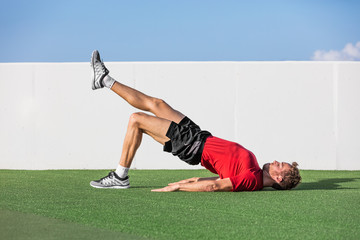 Fitness man doing bodyweight glute single leg floor bridge lift exercises. Fit athlete training glutes muscles with one-legged floor bridge butt raise in summer outdoor gym on grass.