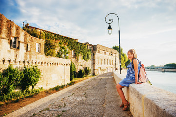 Kid girl tourist resting on quay of Arles, Provence, south of France, wearing blue gingham dress and pink backpack. Travel with children - obrazy, fototapety, plakaty