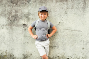 Outdoor portrait of funny little boy standing next to grey wall, wearing cap and backpack
