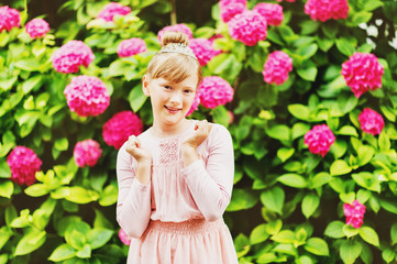 Outdoor portrait of pretty little girl playing with beautiful hydrangea flowers in summer garden, wearing ballerina style dress, hair bun and diamond tiara