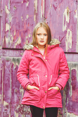 Outdoor vertical portrait of cute 9 year old little girl wearing pink winter coat, standing next to purple background