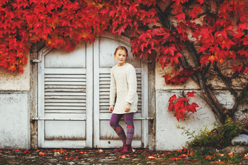 Autumn portrait of pretty little girl wearing knitted beige pullover dress, posing against wall...