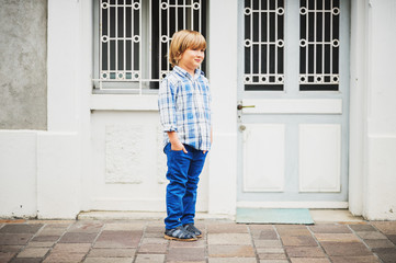 Outdoor fashion portrait of cute little 6 year old boy wearing blue plaid casual shirt and trousers, hands in pockets