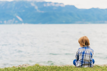 Outdoor portrait of little kid boy playing by the lake on a cloudy day, sitting on the grass, wearing blue shirt. Back view. Image taken at Geneva lake, Lausanne, Switzerland