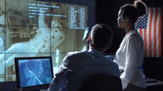 Black and white woman and man monitoring in dark room of space mission control center. Outer works of astronauts. Elements of this image furnished by NASA.