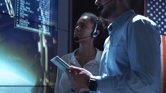 Side view of black woman and white man communicating in Moon mission control center. American flag on background.