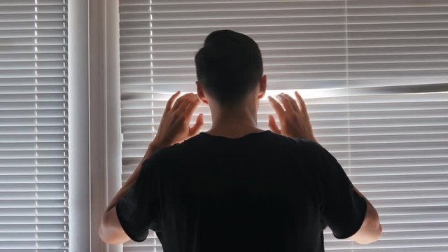 Young man looking out the window through the blinds to the street, spying. suspected