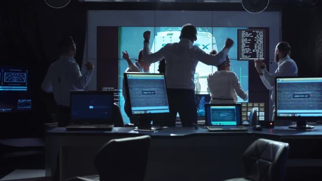 Group of supervisors celebrating docking of spaceship in dark room of mission control center.