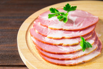 Sliced ham with fresh green lettuce leaves on a round cutting board. Meat products on a brown wooden table.