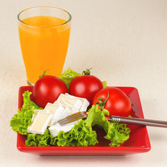 Tomatoes and cheese slices lie on a sheet of fresh salad in a red plate. A glass of orange juice stands side by side on the table.