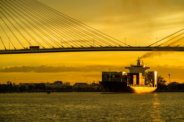 Obraz premium Container cargo ocean ship float on river and pass under bridge at sunset twilight with red sky