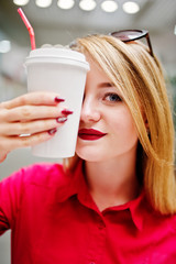 Portrait of a beautiful woman wearing red blouse, casual jeans and black high heels covers half of her face with a cup of coffee in huge shopping mall.