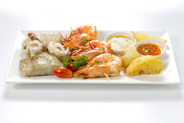 Variation of Roasted Seafood Contain Grilled Jumbo Prawns, Calamari Squids and Barracuda Fish Garlic Pepper with Spicy Chili Sauce, Lemon, Tomatoes, Potatoes, Side View, Selective Focus at Food.