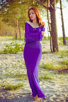 Gorgeous hot young red haired lady at evening sunlight around forest, rocks and water. Woman wearing in purple long dress. Portrait in vintage style.Sweet woman rest on the sea.Nymph on a nature