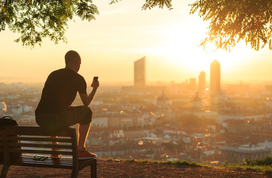 Man on a bench checking his smartphone and enjoying the summer sunrise over a city. Lyon, France.
