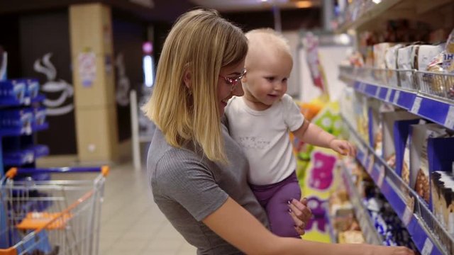 Young mother in glasses holding her child in her arms while choosing cookies on the shelves in the supermarket. Thoughtful mom looking at the product content, carefully choosing products for her child