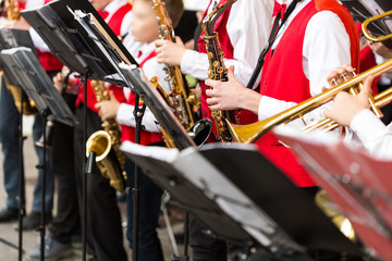 musical instrument, brass band and orchestra concept - closeup ensemble of musicians playing on trumpet and saxophone in red concert costumes, music stand, male hands with equipment, selective focus