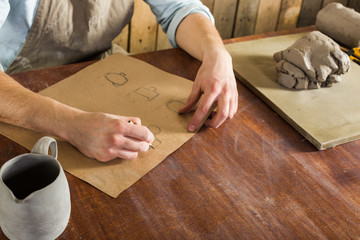 pottery manufacture, artisan tools, ceramics art concept - top view of young man's hands at work on drawing earthenware, a ceramist dressed in a shirt and apron sitting behind desk and finished jug