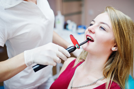 Attractive patient in red-violet dress laying on the dental chair while female dentist treating her teeth with special instruments.