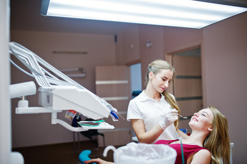Attractive patient in red-violet dress laying on the dental chair while female dentist treating her...