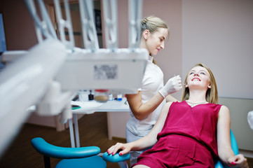 Attractive patient in red-violet dress laying on the dental chair while female dentist treating her...